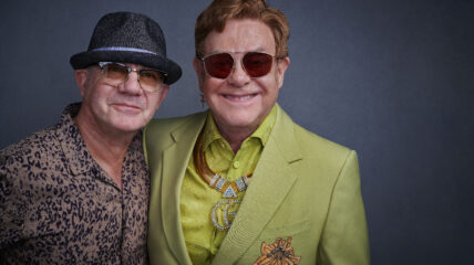 Two white males stand together smiling at the camera. The man on the left is wearing a brimmed brown hat, sun glasses and a brown patterned button down. The man on the right has short brown hair. He's wearing sunglasses and bright green blazer and matching button down shirt.