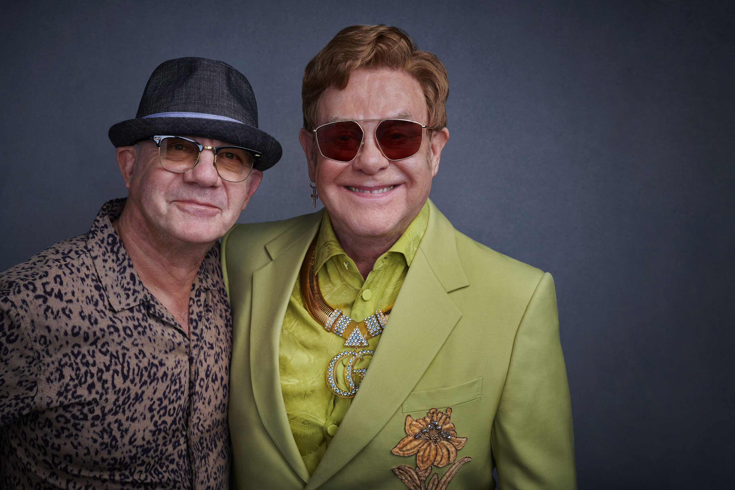 Two white males stand together smiling at the camera. The man on the left is wearing a brimmed brown hat, sun glasses and a brown patterned button down. The man on the right has short brown hair. He's wearing sunglasses and bright green blazer and matching button down shirt.