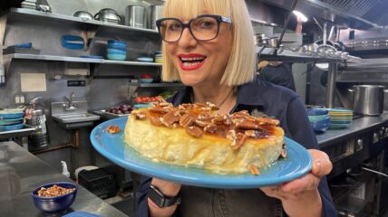 A white woman white with a blonde bob hair cut wearing glasses with Black frames in a restaurant kitchen holds up a cheese cake with pecans on top