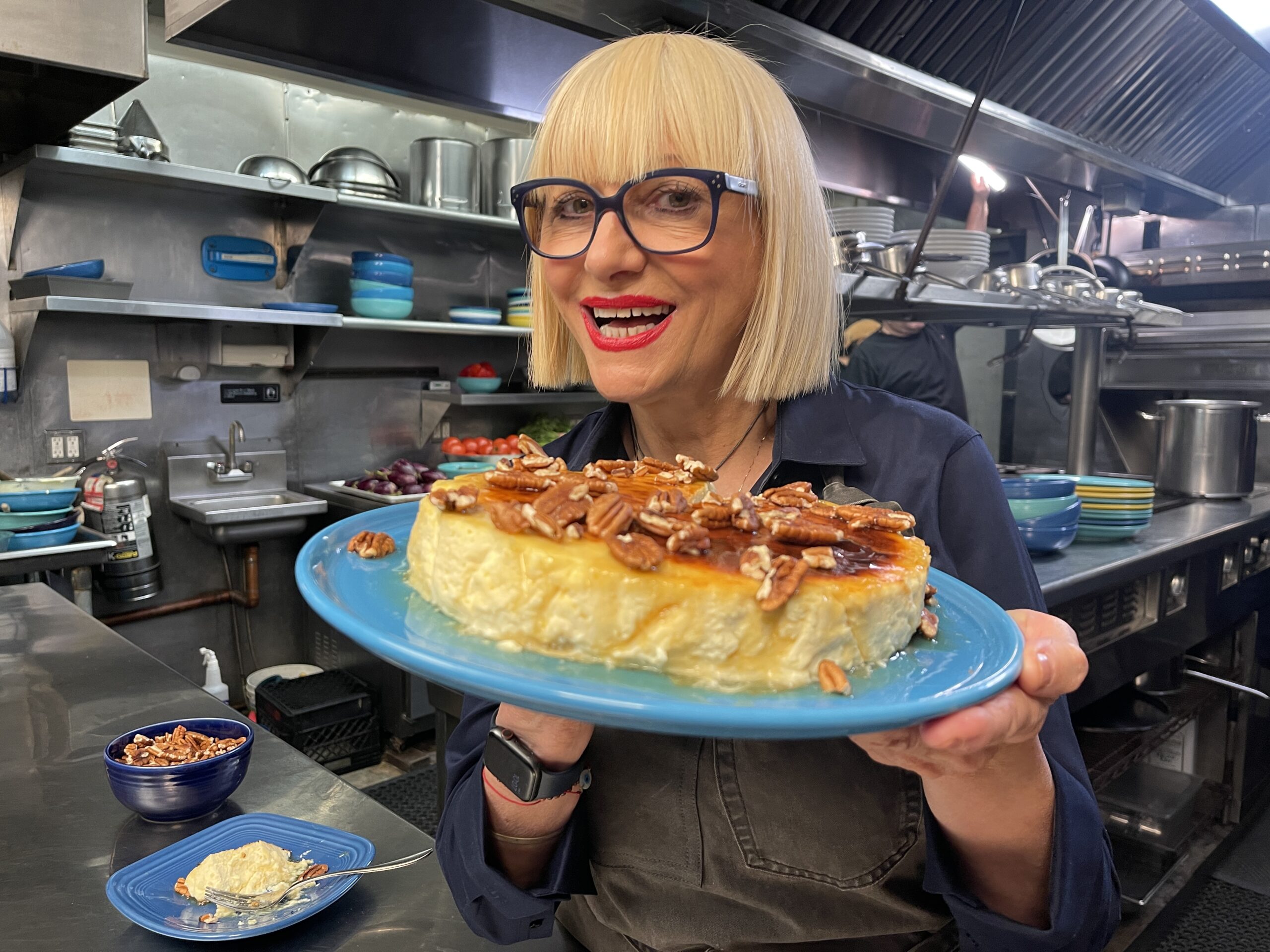 A white woman white with a blonde bob hair cut wearing glasses with Black frames in a restaurant kitchen holds up a cheese cake with pecans on top