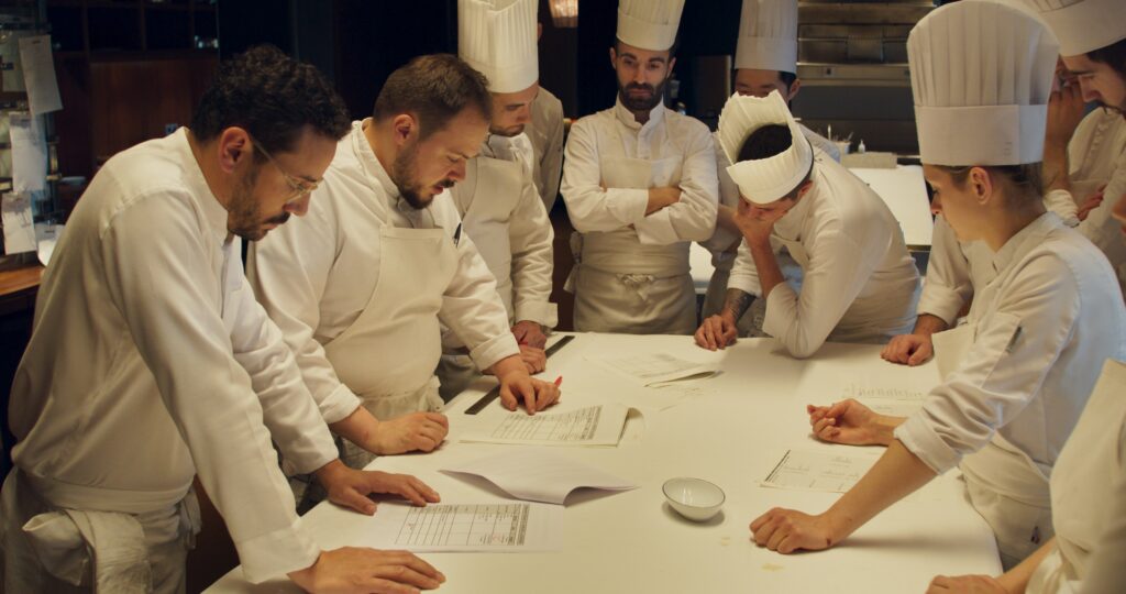 A group of restaurant staff in white chef shirts and aprons standing around a a white counter reviewing papers.