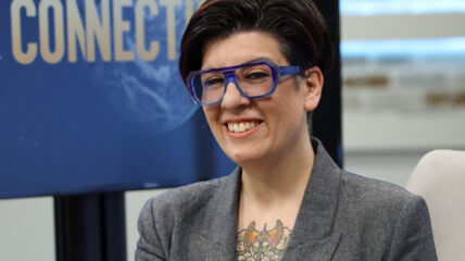 A white woman with dark short brown hair wearing blue framed glasses, a gray tweed blazer and black shirt sits on a TV set with a screen in the background that reads: Environmental Connections