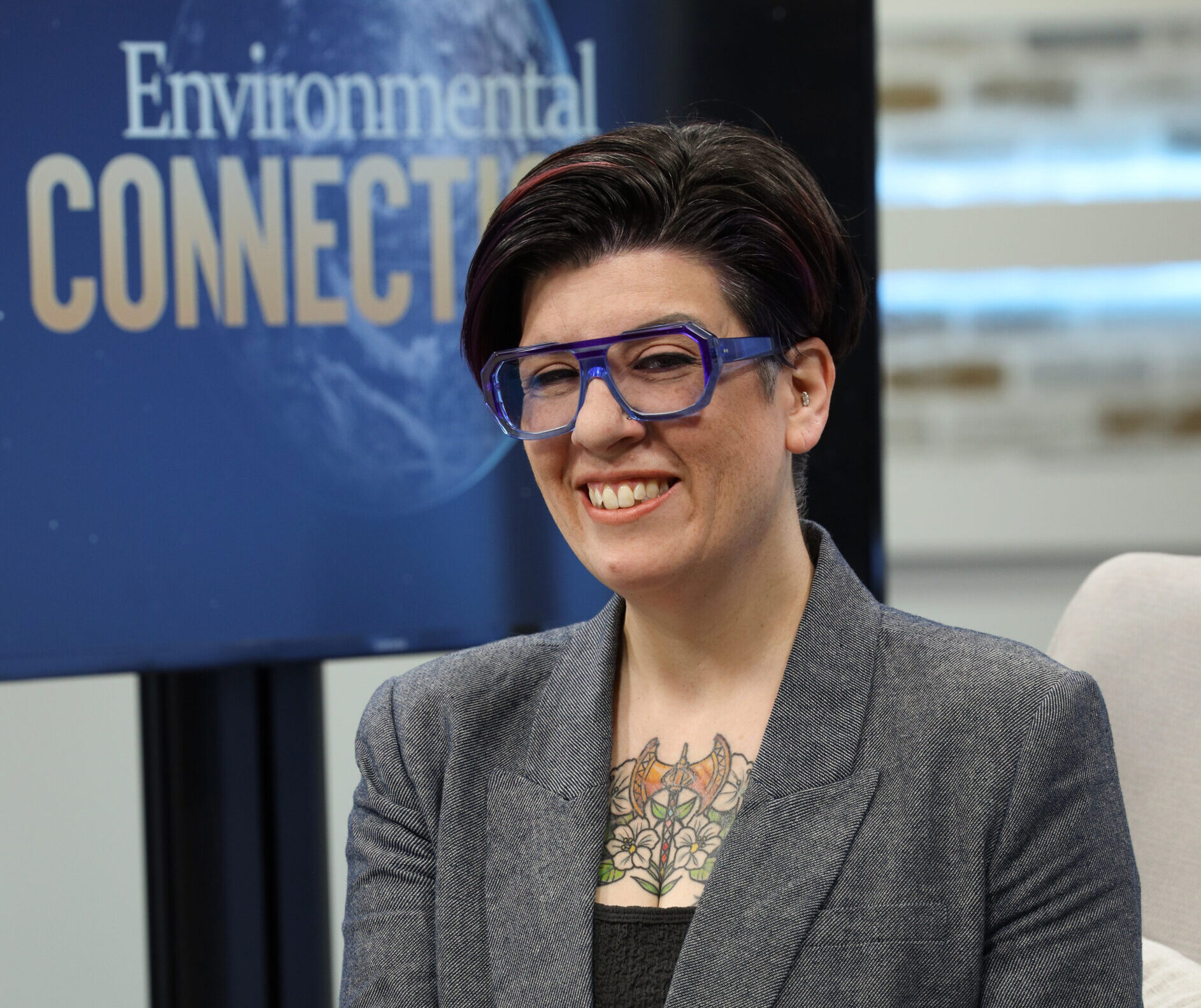 A white woman with dark short brown hair wearing blue framed glasses, a gray tweed blazer and black shirt sits on a TV set with a screen in the background that reads: Environmental Connections