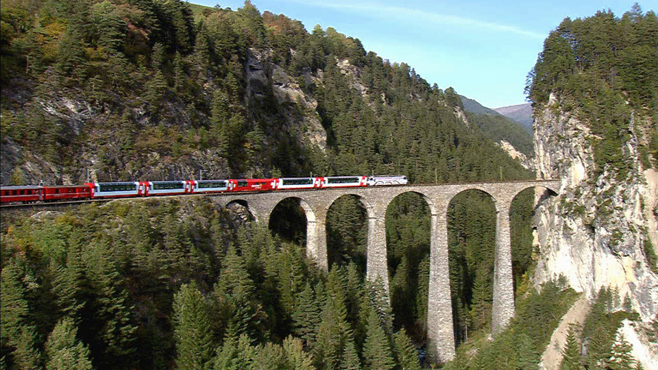 a photo of train on a bridge with treelined mountains in the background
