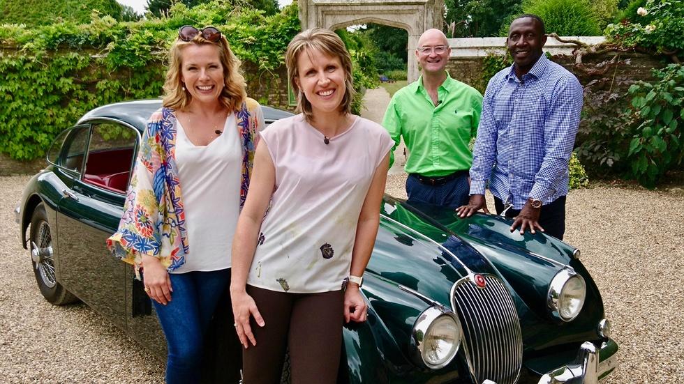 Two white woman both with blonde shoulder length hair wearing white shirts stand in front of a car. A white man in a green shirt and a Black man in the blue shirt stand behind them and the car.