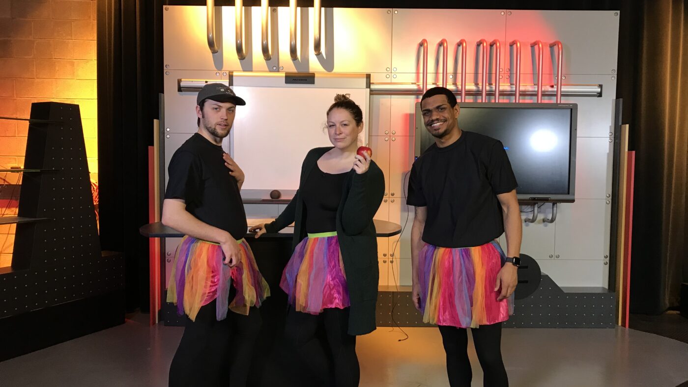 2 men and a woman wearing black leotards and mult-colored tutus in a TV studio