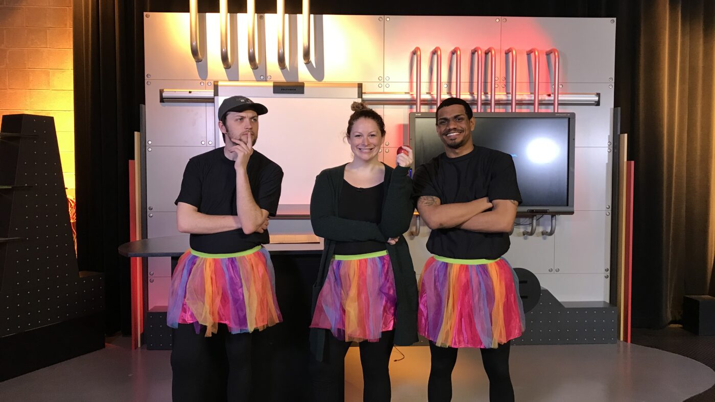 Two men and a woman are wearing black leotards and tutus in a TV studio