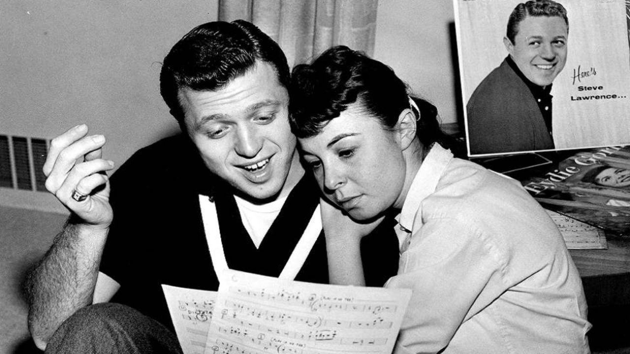 A Black and white photo of a white man with dark short hair wearing a dark shirt and a white woman with dark hair pulled back wearing a white blouse. She is resting her check on his shoulder as the two look over a sheet of music.