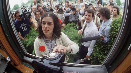 A white woman with brown shoulder length hair wearing a white cardigan stands in front of a window with her hand on a Black rotary phone. Behind her is a bunch of press people and cameras