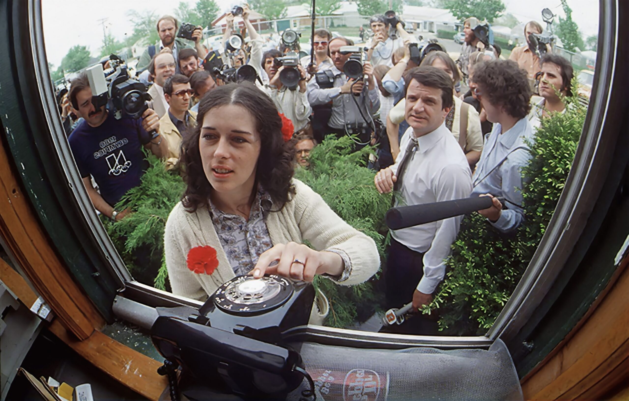 A white woman with brown shoulder length hair wearing a white cardigan stands in front of a window with her hand on a Black rotary phone. Behind her is a bunch of press people and cameras