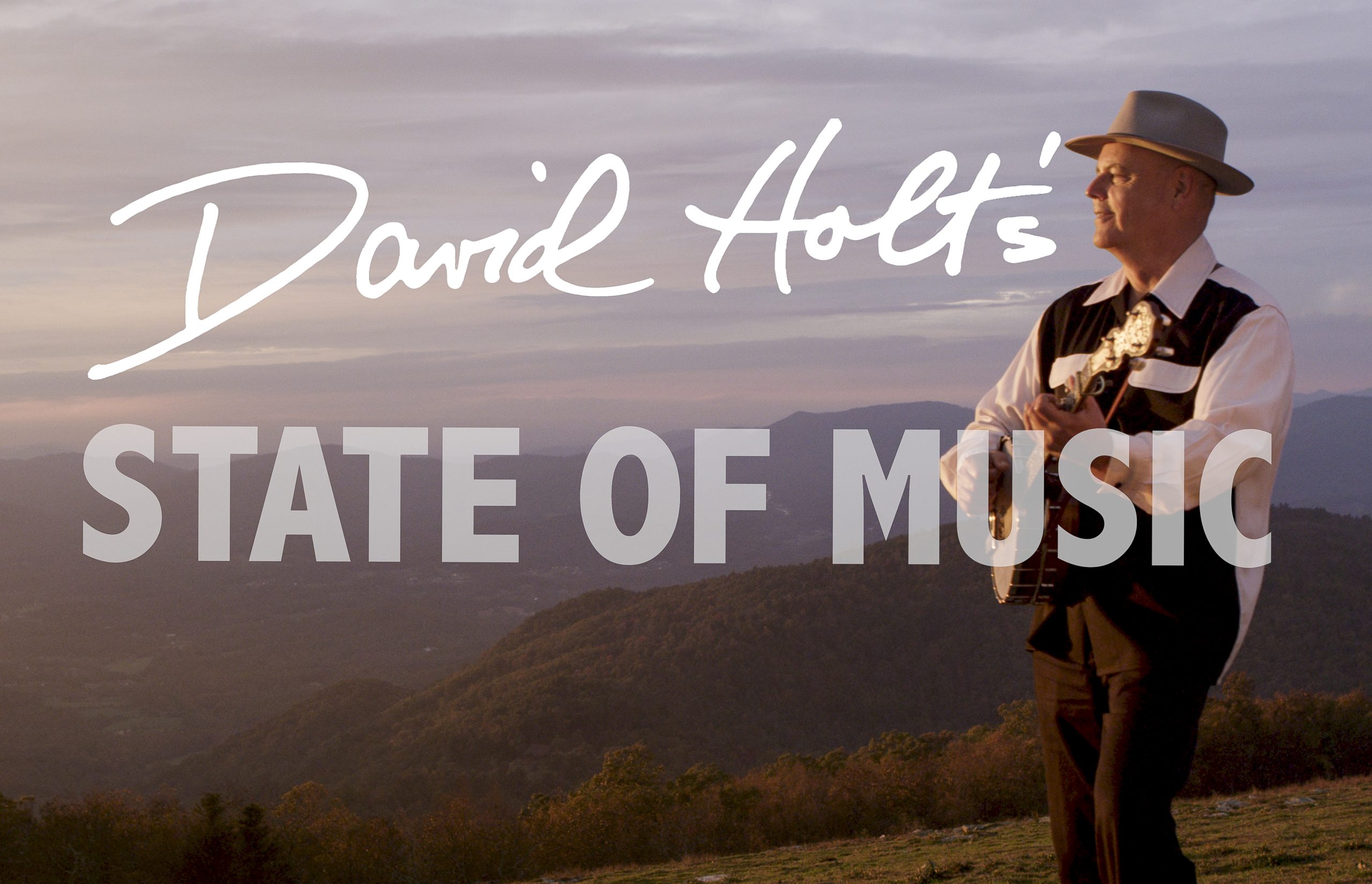 A white male wearing a brown hat, white and brown shirt and brown pants playing guitar outside with mountains in the background.