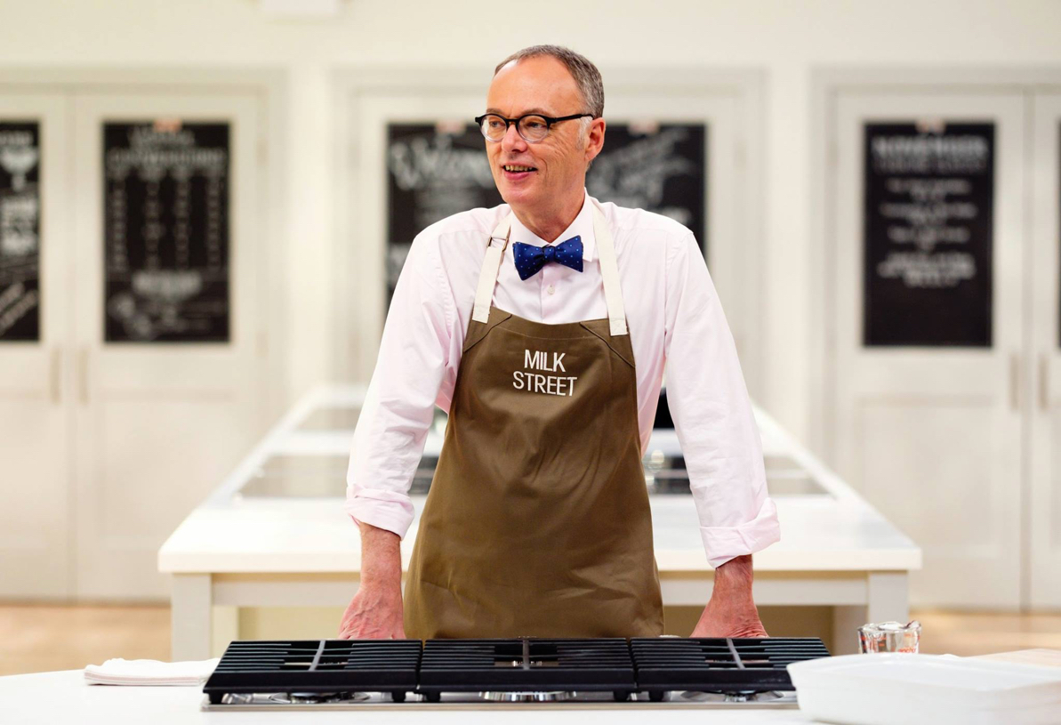 A white man with short gray hair wearing round framed glasses, a white collared shirt, blue tie and a brown apron stands in front of a cooking range.
