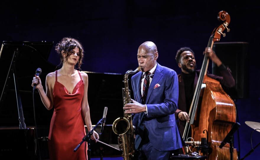 A Black musician plays the sax, wearing a blue suite with a white shirt. A white woman in a red dress holds a microphone watches him. A Black man playing the upright bass stands behind him.