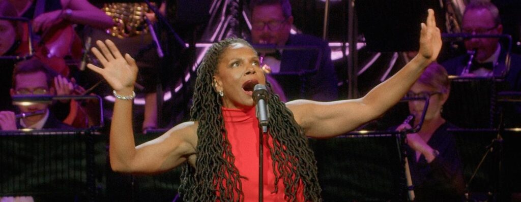 A Black woman with long wavy Black hair wearing a red sleeveless dress has her arms raised as she sings into a microphone