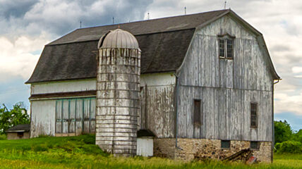 A white barn with a dark brown roof and a shite silo on a farm