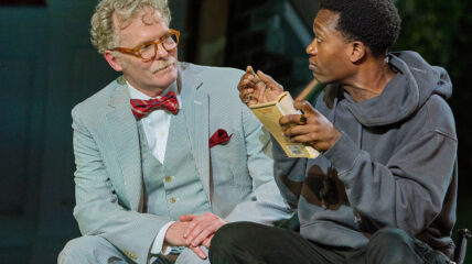 Am older white man with gray hair in a gray 3-piece suite with a red bow tie sits next to a young Black man wearing a gray hoodie sweatshirt holding a paper back book.