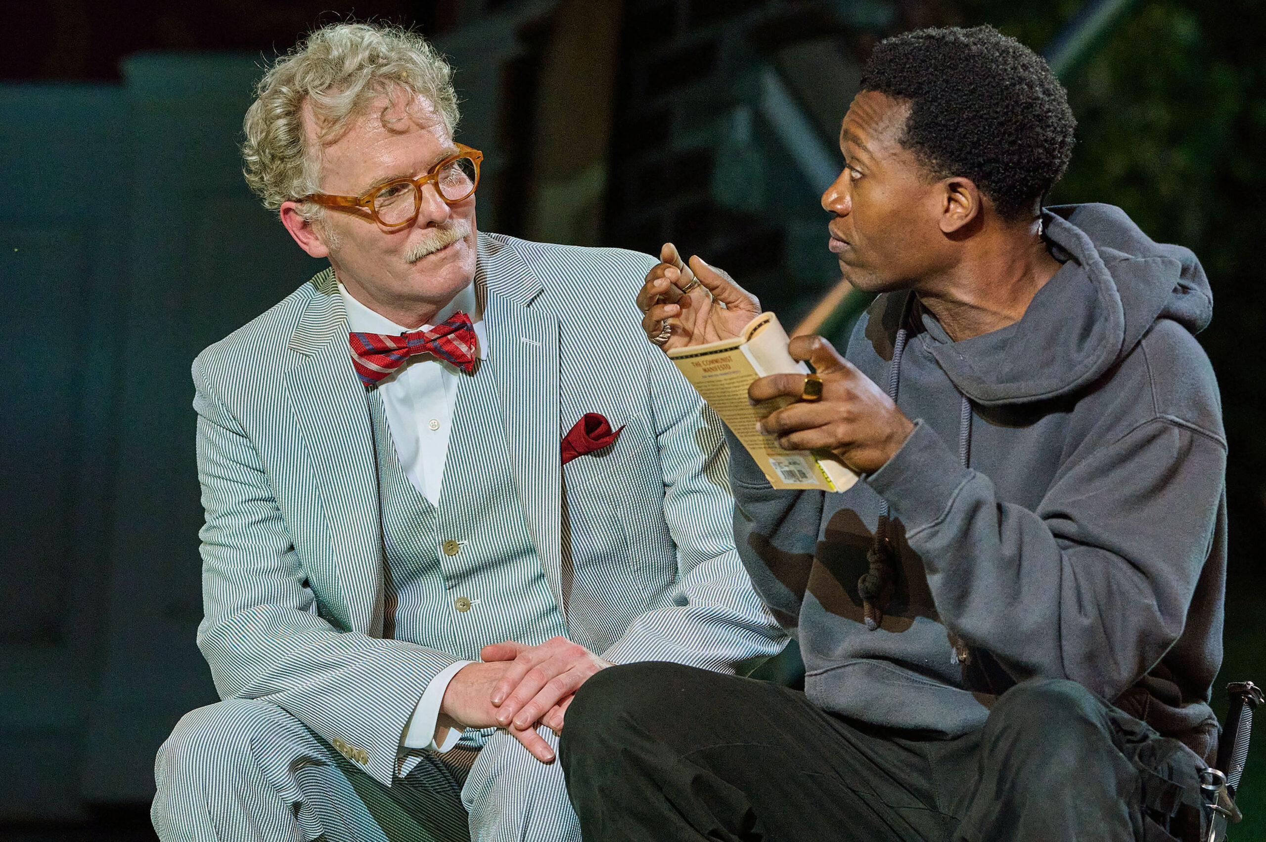 Am older white man with gray hair in a gray 3-piece suite with a red bow tie sits next to a young Black man wearing a gray hoodie sweatshirt holding a paper back book.