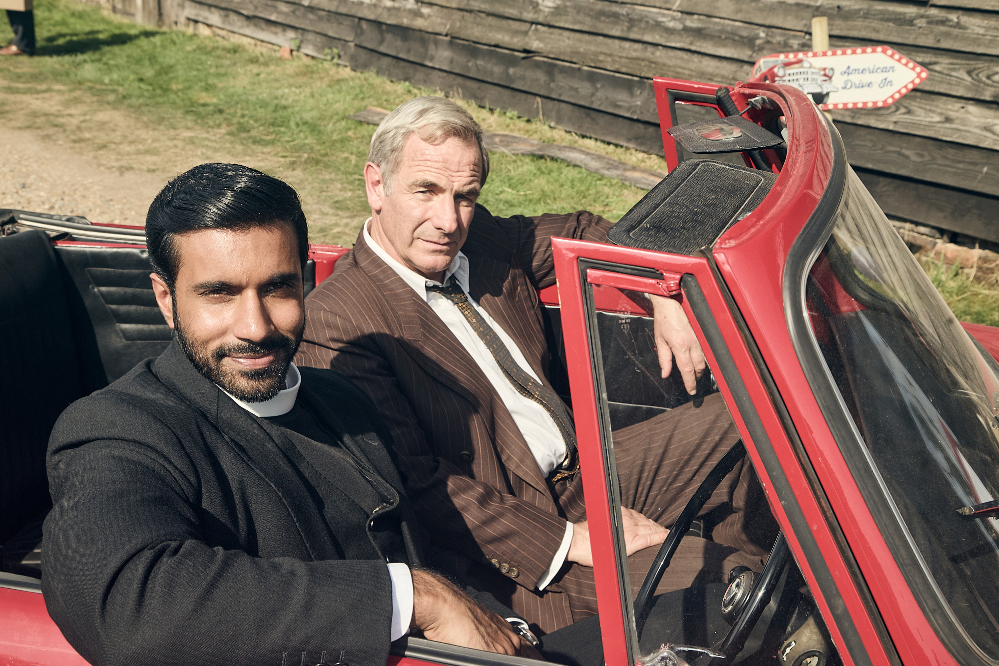 An older male wearing a brown suit sits next two a young vicar with black hair and a beard wearing all black. They are in a red convertible car.