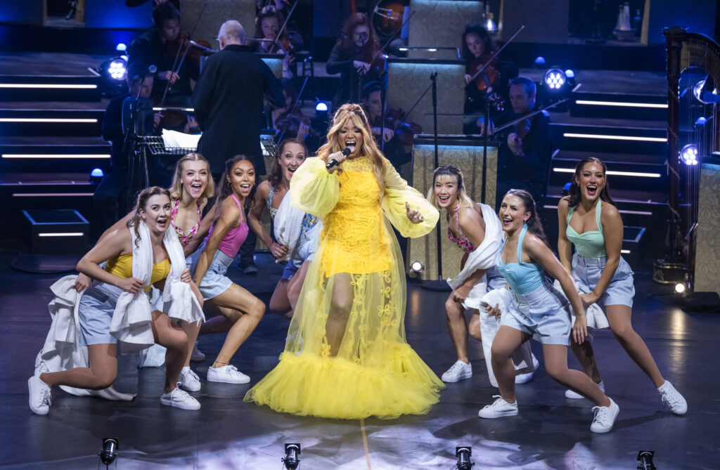 A Black female with long blonde hair wearing a yellow gown and singing into a microphone with six female dancers circling around her.