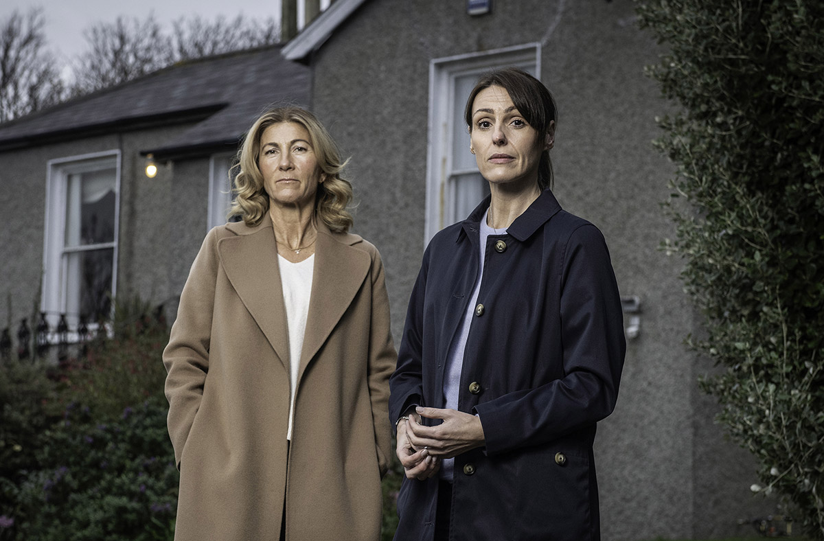 Two woman stand side by side. On the left is a white woman with blonde hair wearing a light brown coat. On the right, a woman with brown hair pulled back wearing a blue coat