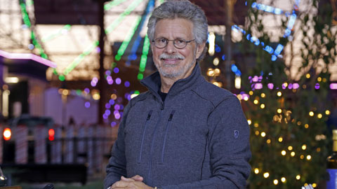 A white male with gray hair, mustache and beard with round, Black framed glasses wearing a blue seater smiles at the camera