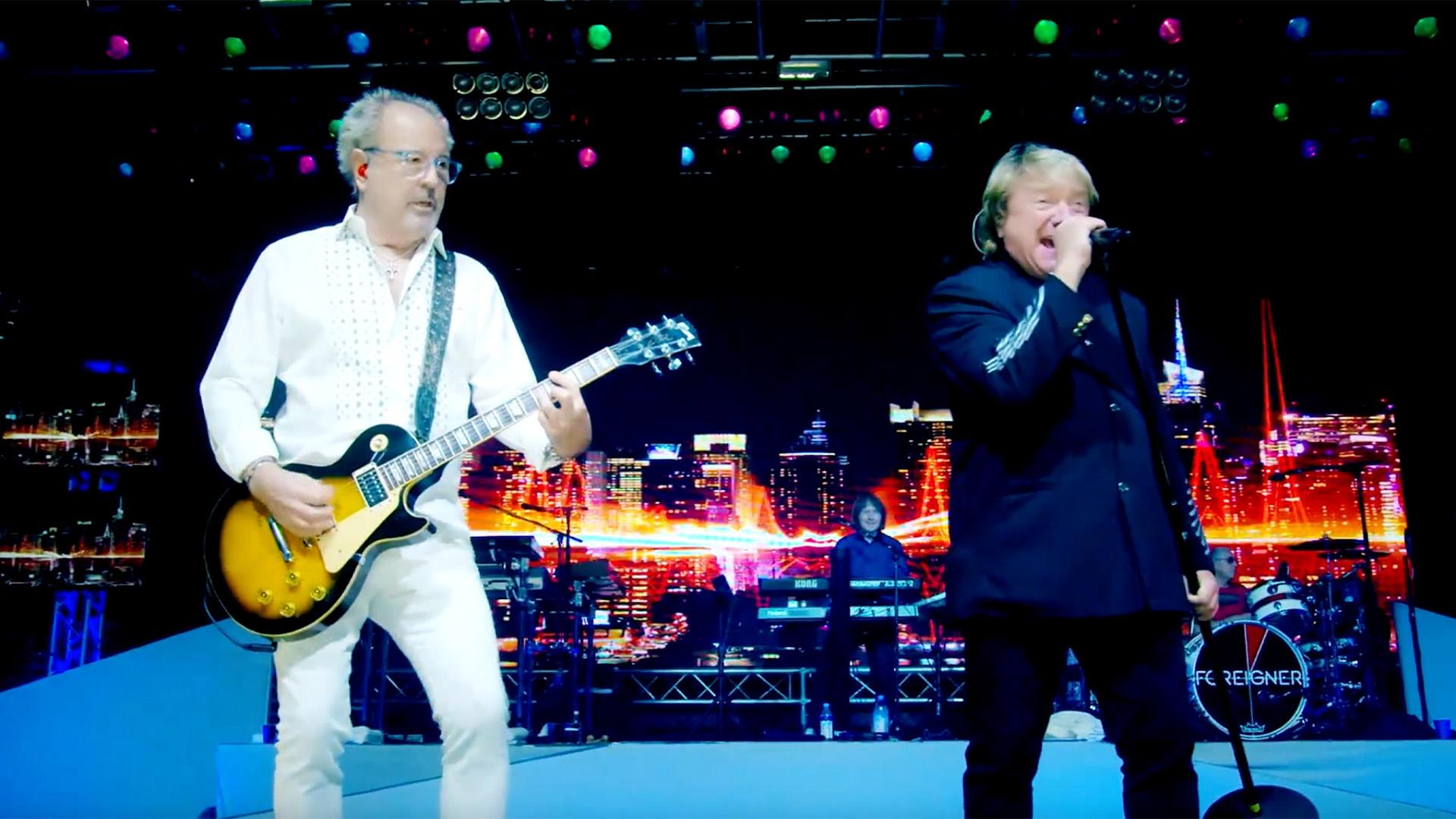 Two white men on stage. One is wearing all white playing the guitar. The other wearing all Black is singing.