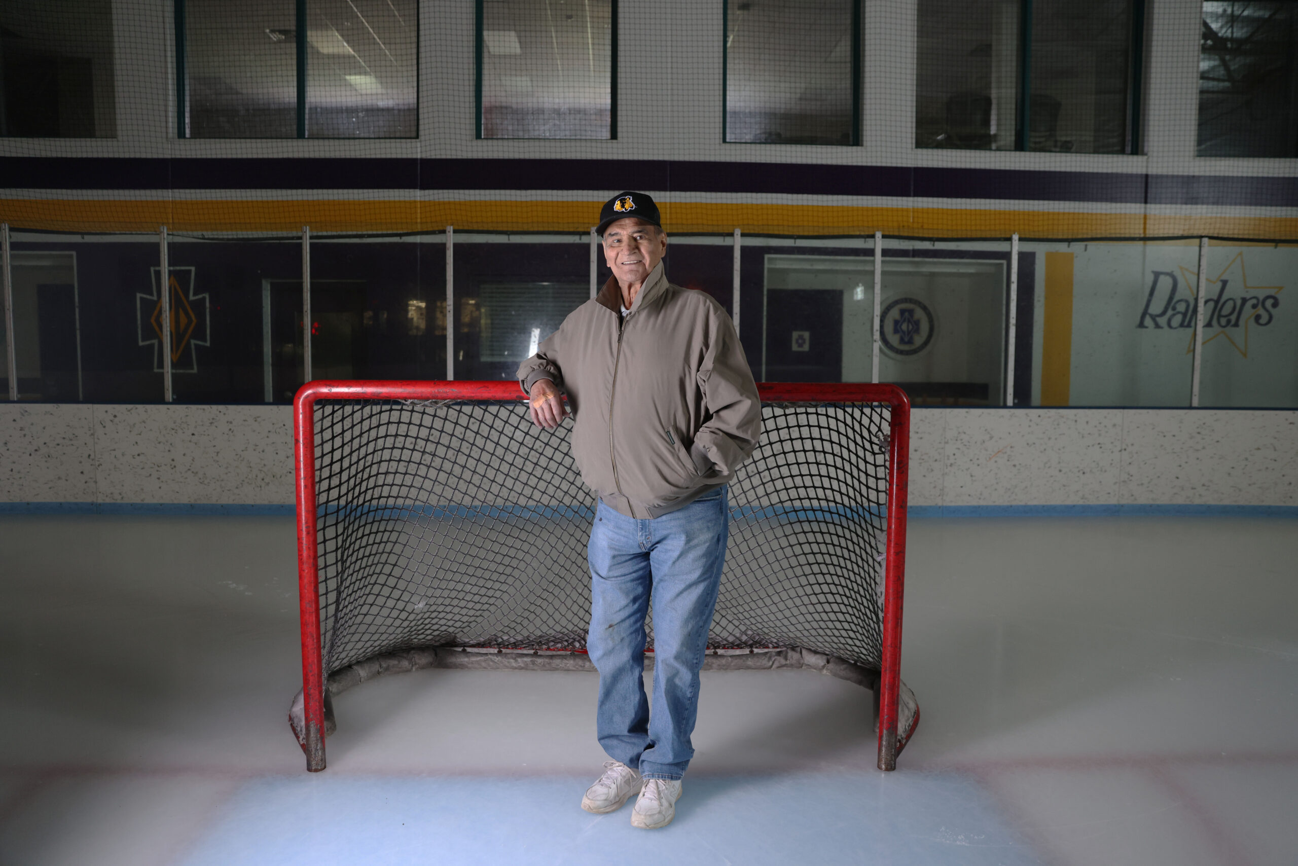 Henry Boucha wearing a tan coat, jeans and a baseball cop standing in front of a goal on an ice rink.