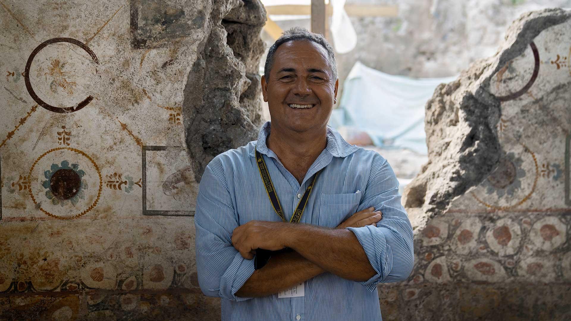 An archaeologist, a white male with short gray hair wearing a blue button-down shirt with his hands folded smiles.
