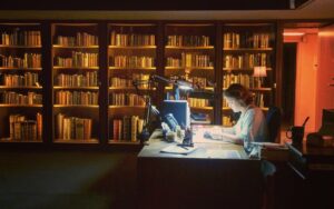 a librarian sits at a desk with a wall of books behind her.