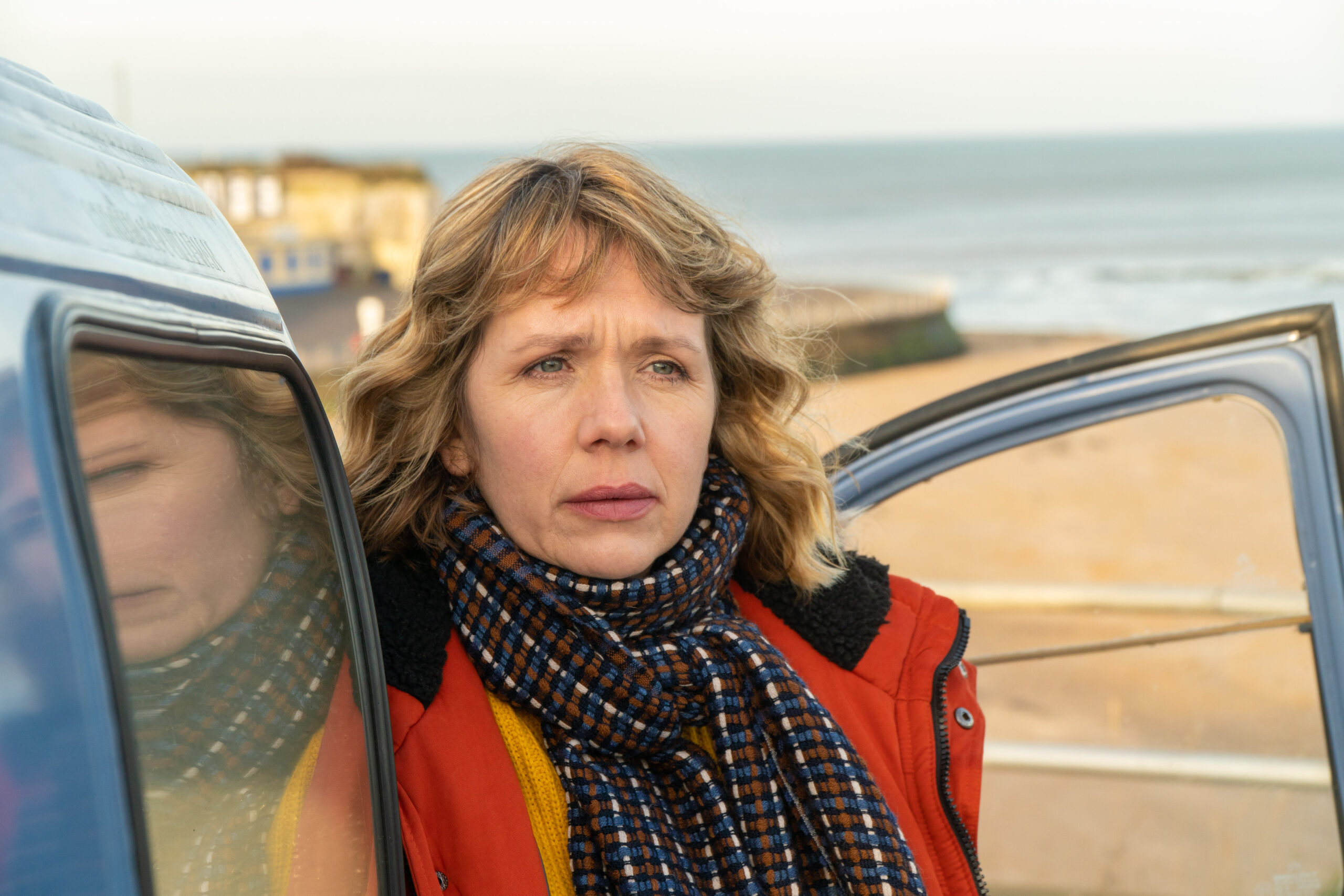 A blonde woman with a colorful scarf and red coat stands in front of an open door to a car.
