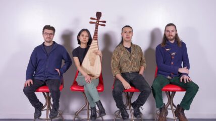 Four band members sit on a row of red chairs