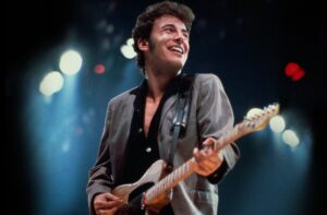 A young Bruce Springsteen wearing a gray blazer and Black button down playing guitar on stage.