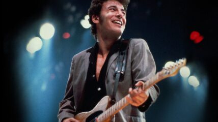 A young Bruce Springsteen wearing a gray blazer and Black button down playing guitar on stage.