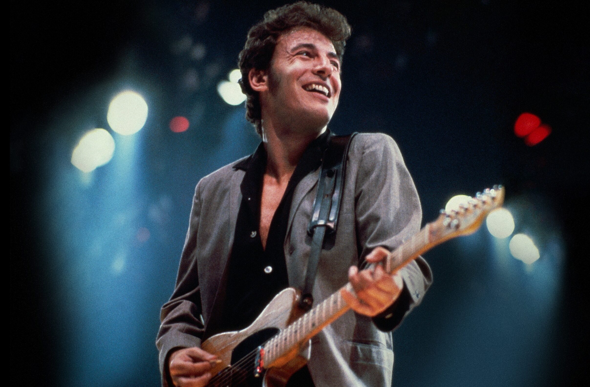 A young Bruce Springsteen wearing a gray blazer and Black button down playing guitar on stage.