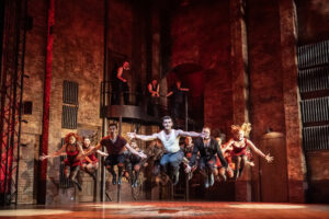 A white man in a white t-shirt and jean is dancing on stage. Other men and women are dancing beside him.