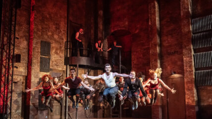A white man in a white t-shirt and jean is dancing on stage. Other men and women are dancing beside him.