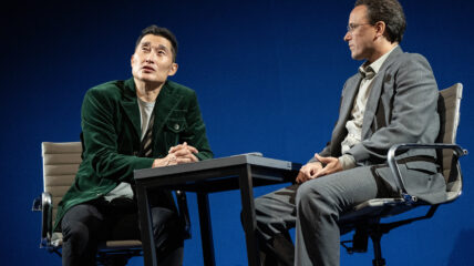 An Asian man in a dark suite sits at a table with a white man in a gray suit talking.