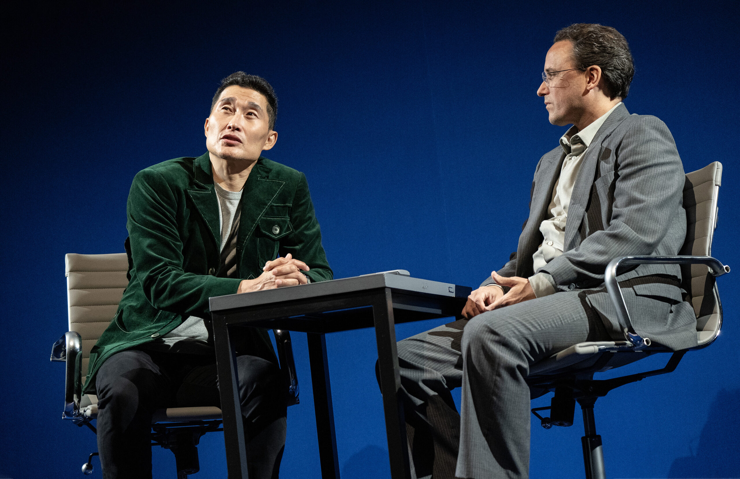 An Asian man in a dark suite sits at a table with a white man in a gray suit talking.