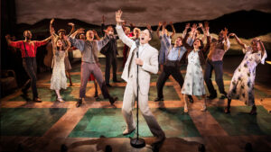 A white man in a tan suite and white shirt and tie stands on stage singing with one hand raised in the air. People are surrounding him.