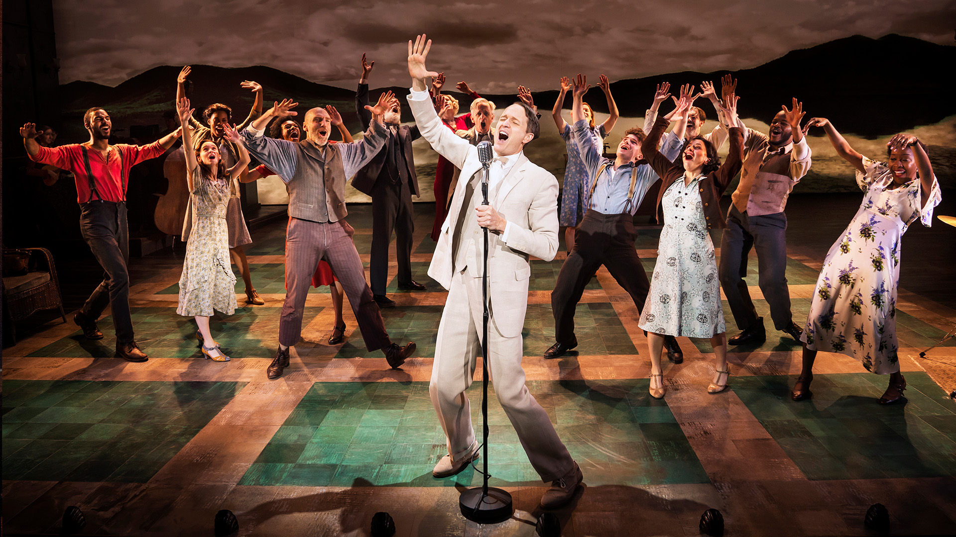A white man in a tan suite and white shirt and tie stands on stage singing with one hand raised in the air. People are surrounding him.