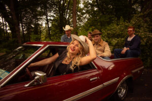 A blonde woman in a Black leather vest and white cow boy hat drives a maroon car, with four men sitting in the open back.
