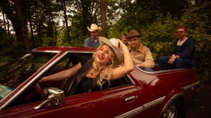 A blonde woman in a Black leather vest and white cow boy hat drives a maroon car, with four men sitting in the open back.