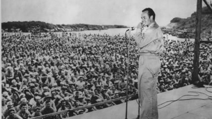 Bob Hope stand on stage in front of crowd of soldiers