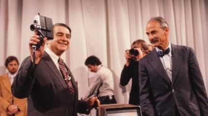 A white man in a gray suit holding up a polaroid camera. Another man also wearing a gray suit and tie stands next to him staring at him.