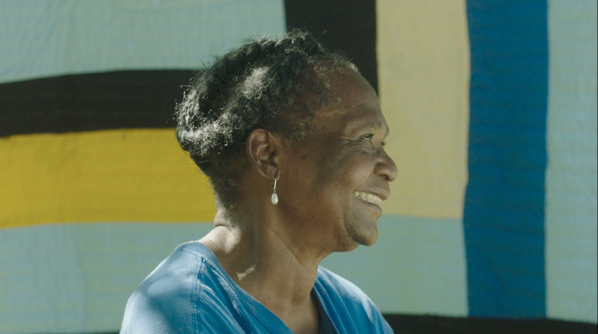 An older Black women with Black hair pinned backed wearing a blue blouse smiles. In the background is a quilt.