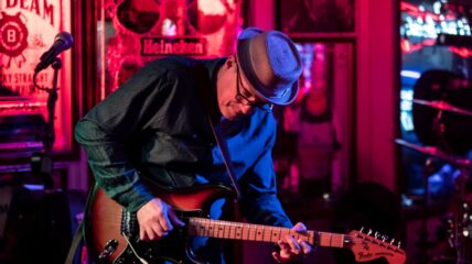 A white male wearing a dark shirt and hat playing the electric guitar.