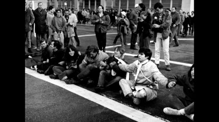 A row of teen men and women sitting along a road holding hands in protest of the Vietnam War.