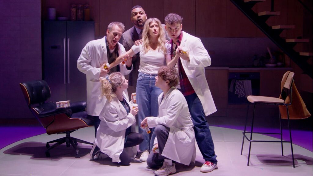 A young white girls with long blonde hair wearing a white t-shirt and jeans is surrounded by doctors in white lab oats.