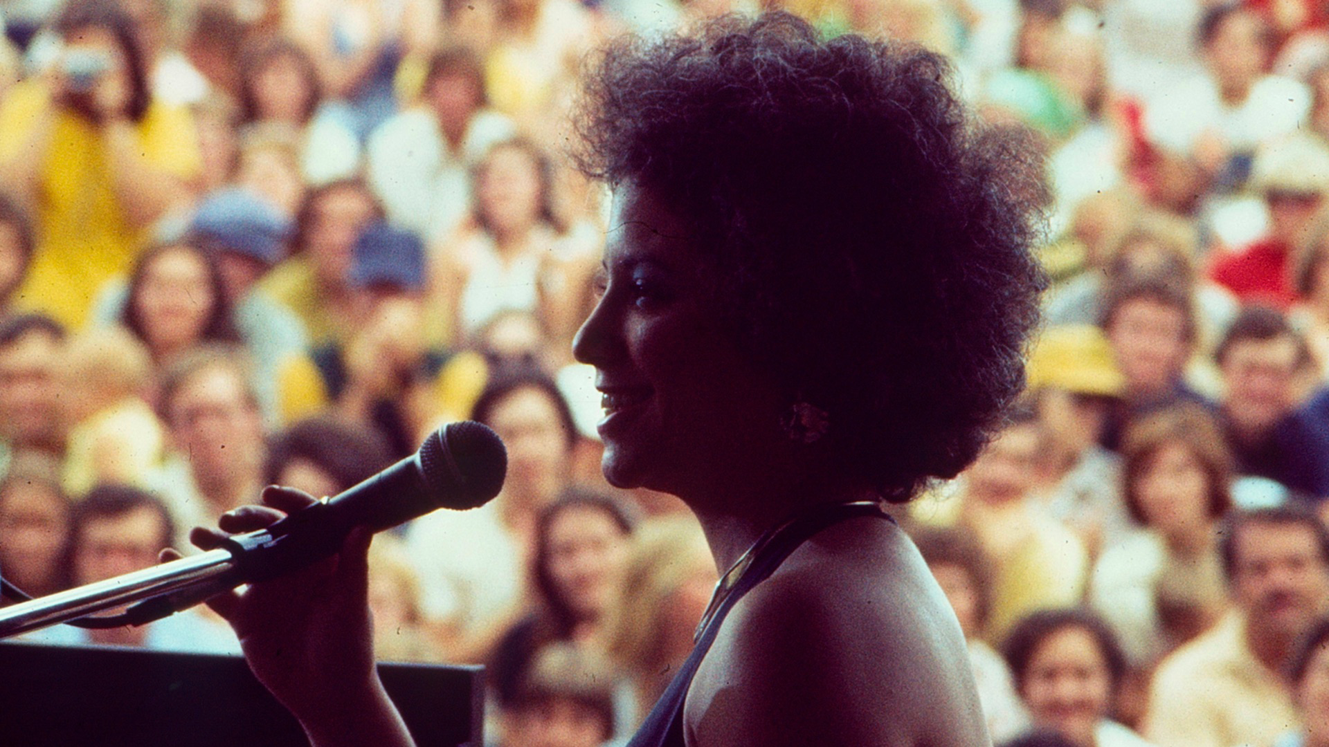 A woman with dark hair smiling with a mic in front of a large crowd.