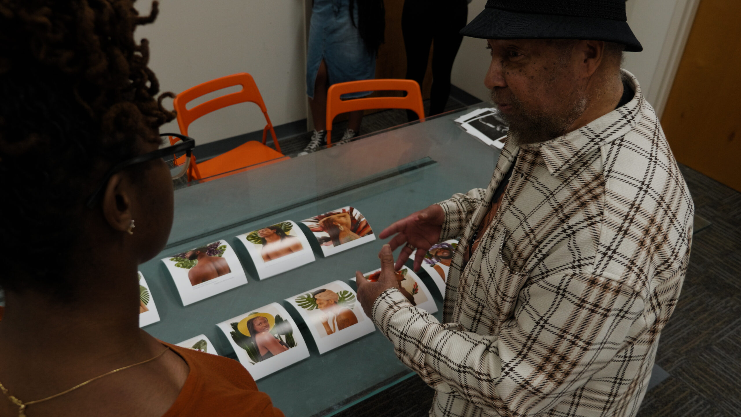 An older Black man talking to a younger Black woman as they stand close to a table full of portrait-style photographs.